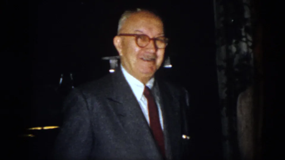 In 1958 Virginia home-movie footage, an older man in glasses, suit, and red tie smiles during a nighttime gathering.