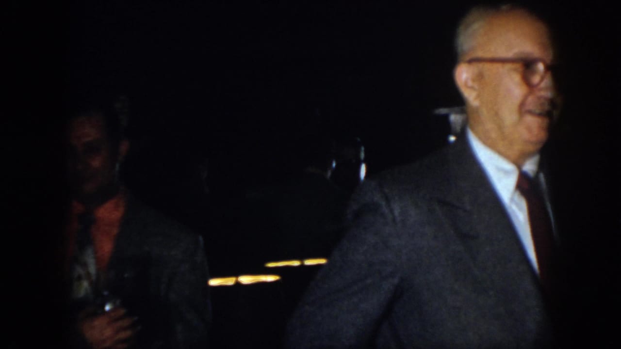 Older suited man turns at right while another man stands in shadow.