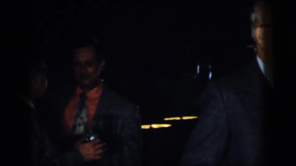 A man in a jacket holds a drink in a dark nighttime gathering, with partial suited figures around him.
