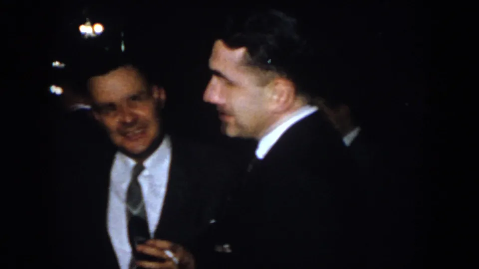 Two well-dressed men in suits converse at a 1958 Virginia nighttime social gathering, one holding a drink glass.