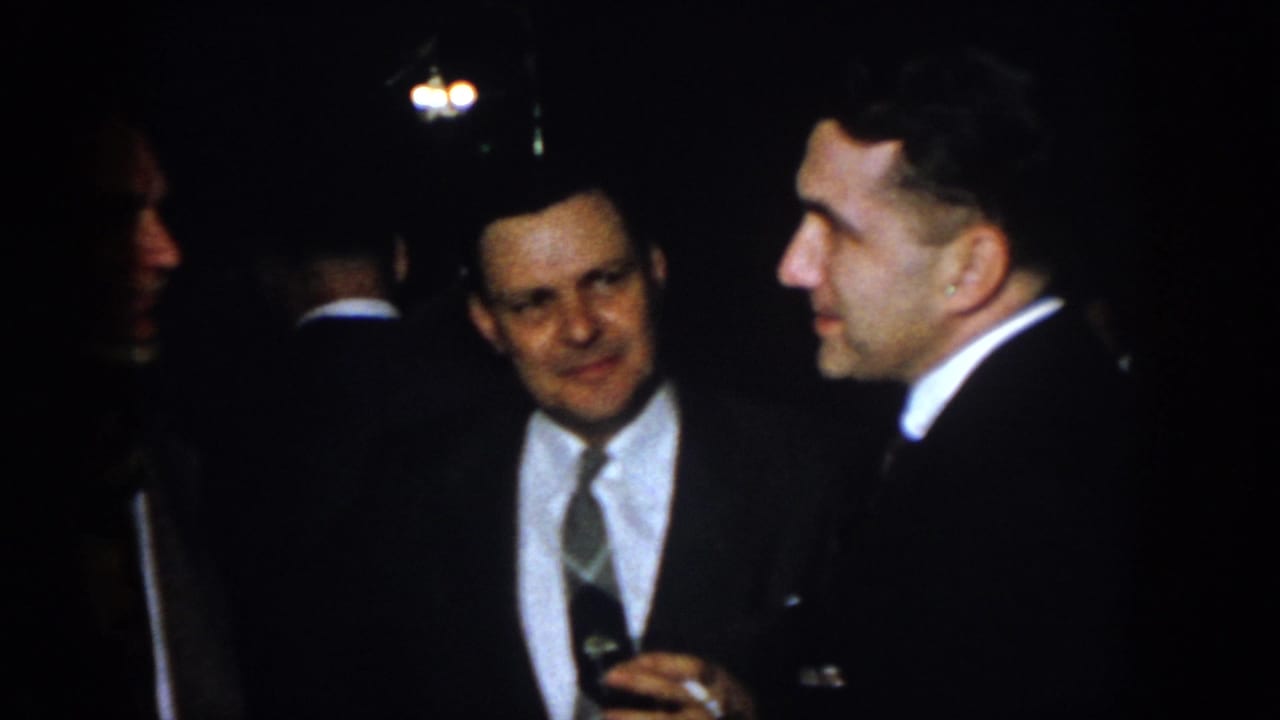 1958 Virginia home-movie frame showing suited adult men talking in low light, including a man with glasses holding a drink.