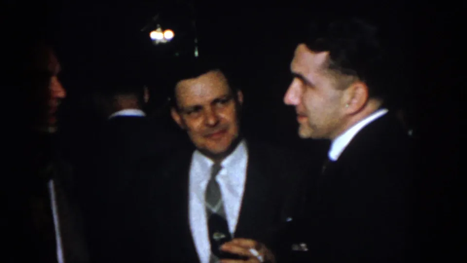 1958 Virginia home-movie frame showing suited adult men talking in low light, including a man with glasses holding a drink.