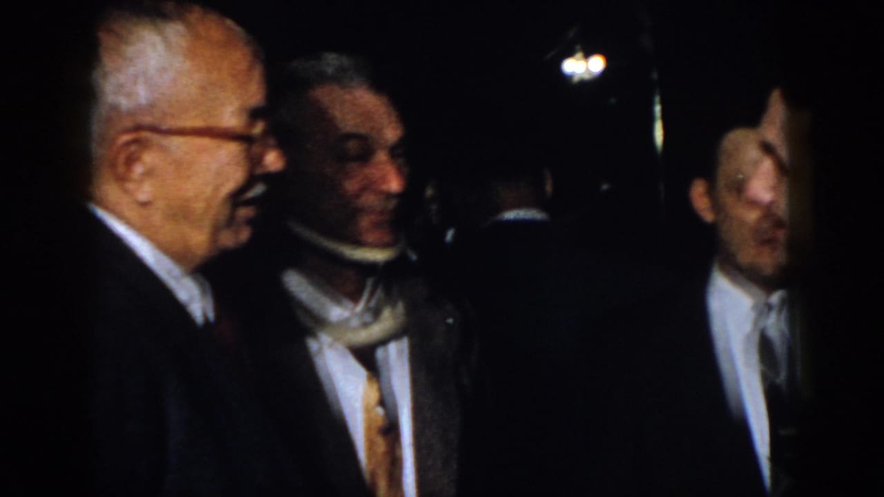 Older man with glasses stands among other men in suits in a dark room.