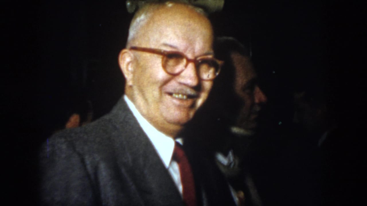 1958 Virginia home-movie frame showing an older suited man with glasses smiling near other men indoors.