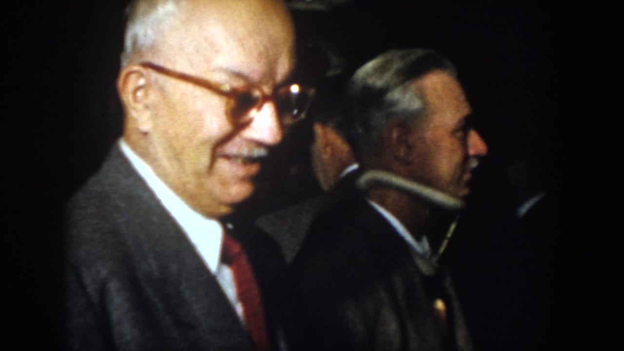 Older man in glasses turns while smiling, with another suited man in profile behind him.