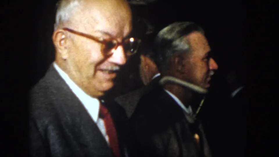 1958 Virginia frame of a smiling older man in a suit and glasses beside other men in a dark indoor space.