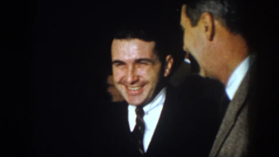 1958 Virginia home-movie frame showing a smiling younger man in a dark suit among other men indoors.