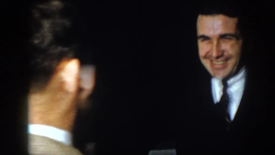 1958 Virginia frame with a smiling younger suited man partly separated by foreground figures in a dark interior.