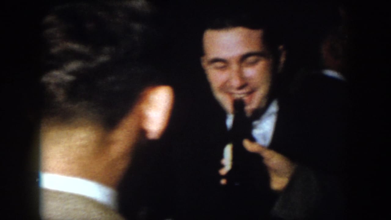 1958 Virginia home-movie frame of adult men in suits gathered indoors, one holding a bottle near another smiling man.