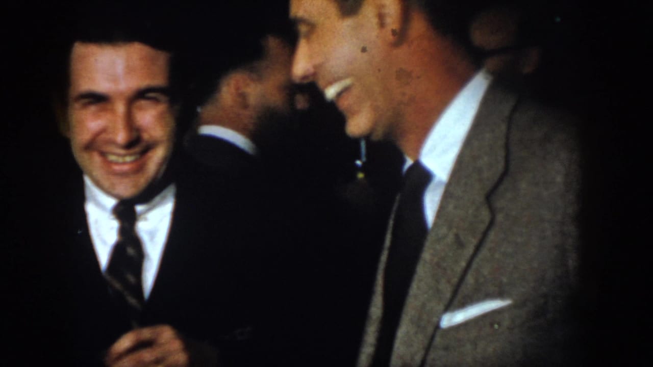 1958 Virginia frame showing a smiling suited man looking toward the camera during an indoor social gathering.