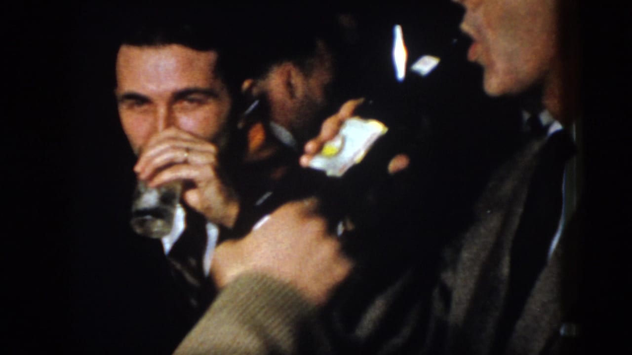 Man in a suit drinks from a glass while others stand nearby indoors.