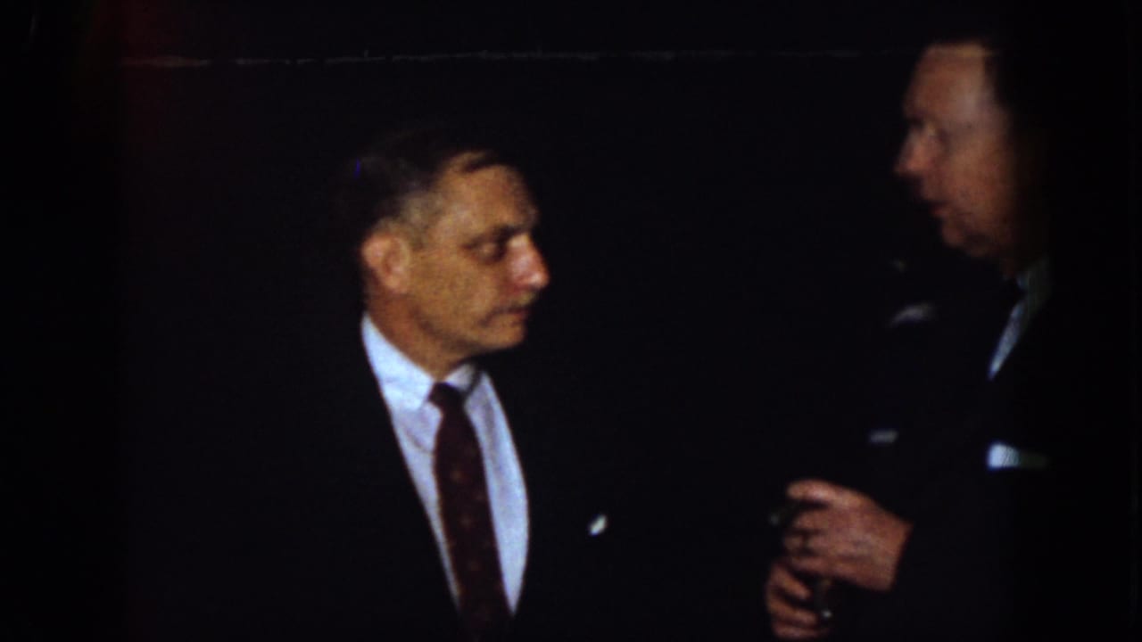 1958 Virginia home-movie frame showing suited men in low light during a formal conversation, with one man holding a drink.
