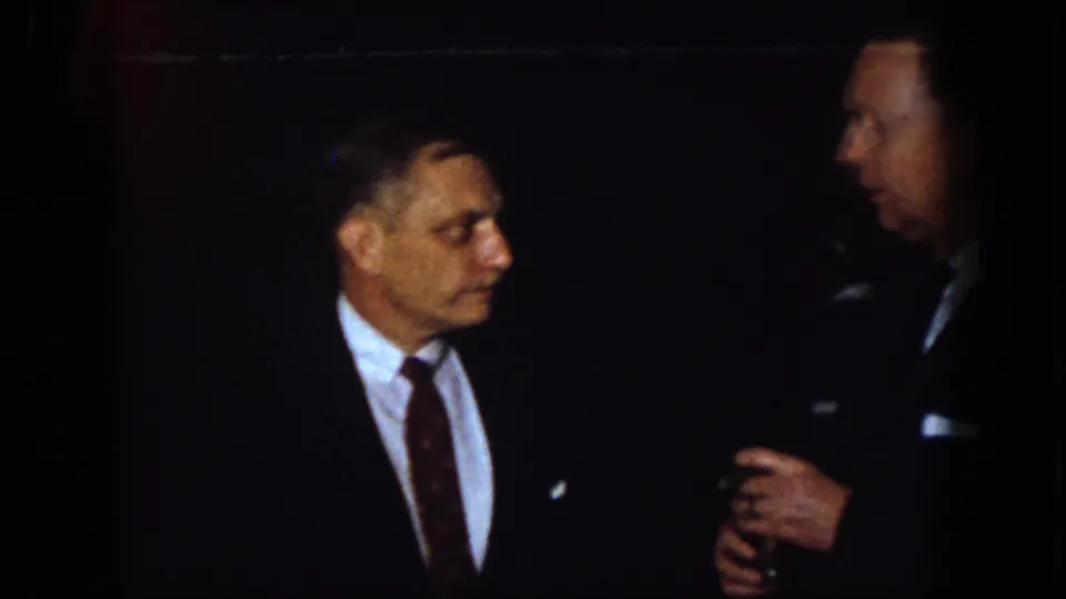 1958 Virginia home-movie frame showing suited men in low light during a formal conversation, with one man holding a drink.