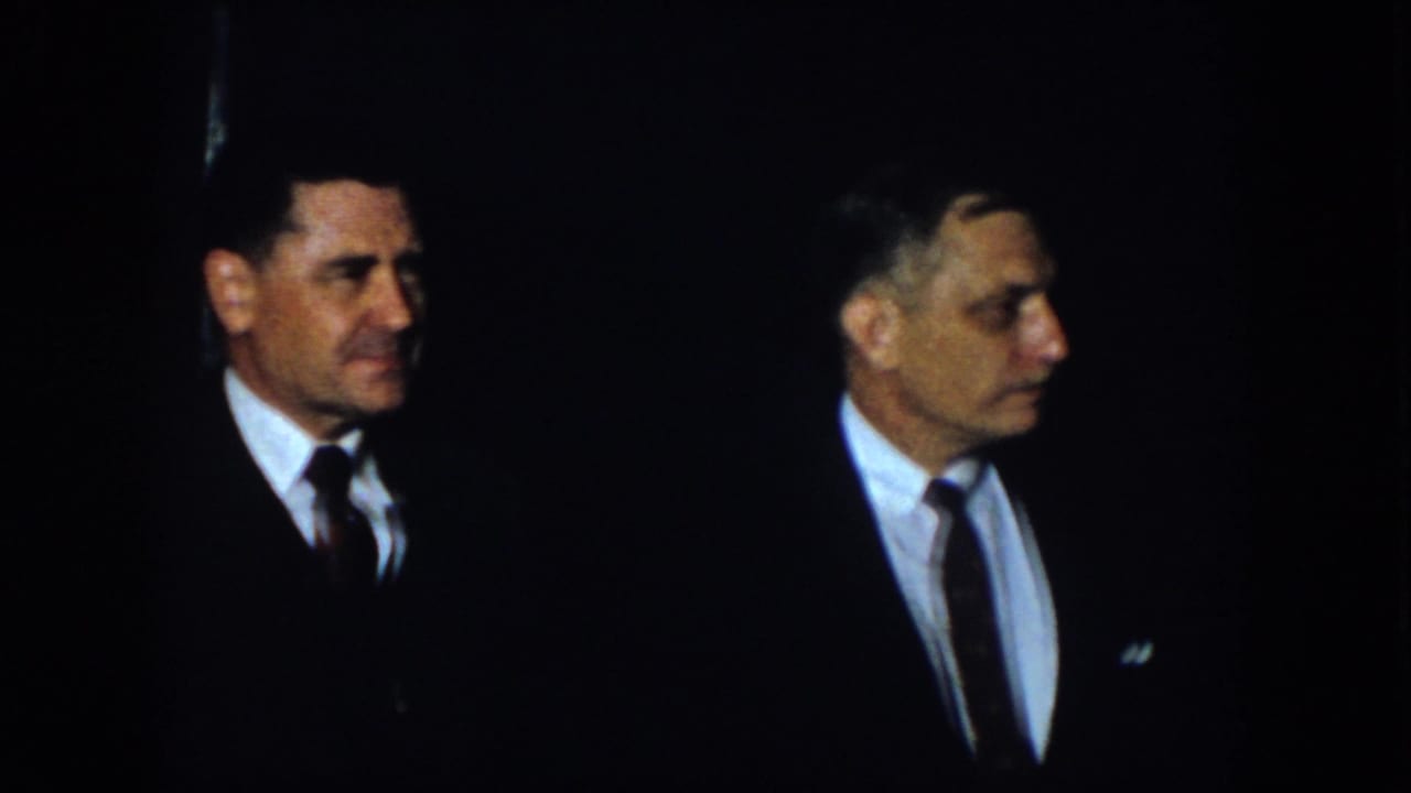 Two men in dark suits face each other against a mostly black background.
