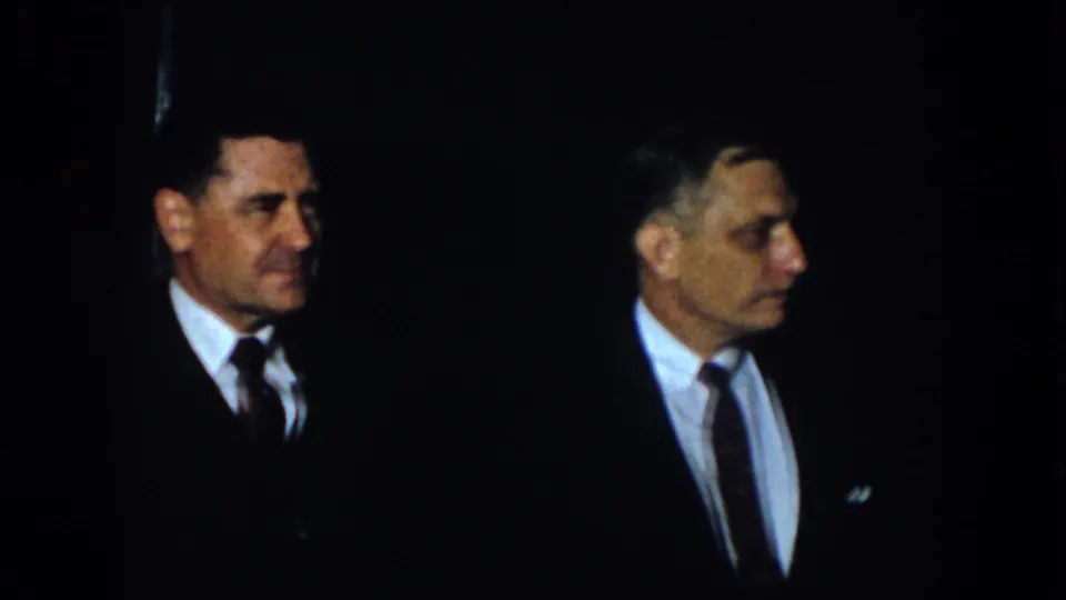 1958 Virginia frame of two suited adult men facing one another in a dim indoor setting with strong shadow around them.