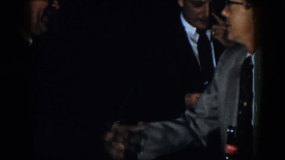 1958 Virginia frame showing suited men in a dim room during a close greeting, with hands visible near the center foreground.