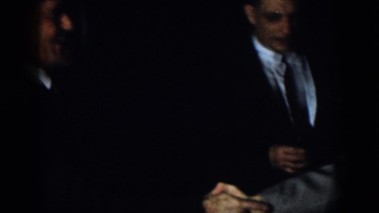 Virginia 1958 home-movie frame showing formally dressed men seated in a very dark interior, including a partly blurred man in the foreground and hands visible near the lower edge.