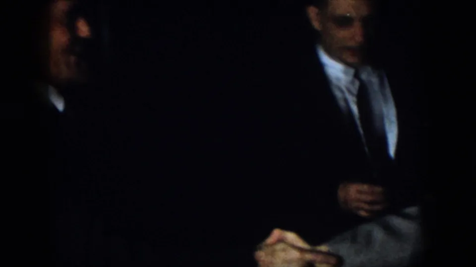 Virginia 1958 home-movie frame showing formally dressed men seated in a very dark interior, including a partly blurred man in the foreground and hands visible near the lower edge.