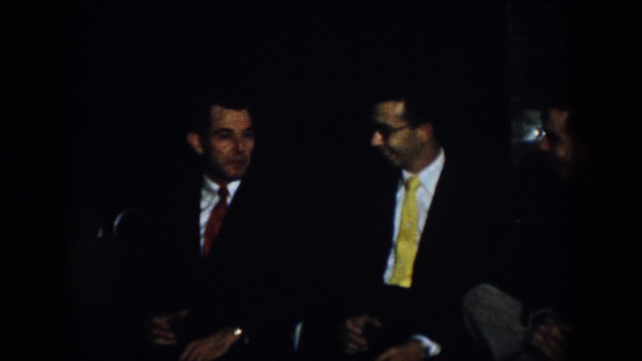 Two men in suits and ties sit and talk in a dark interior.