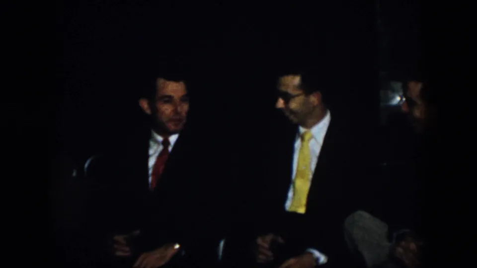 Virginia 1958 home-movie frame showing two seated men in dark suits, one wearing a red tie and another wearing eyeglasses with a yellow tie, conversing in low light.