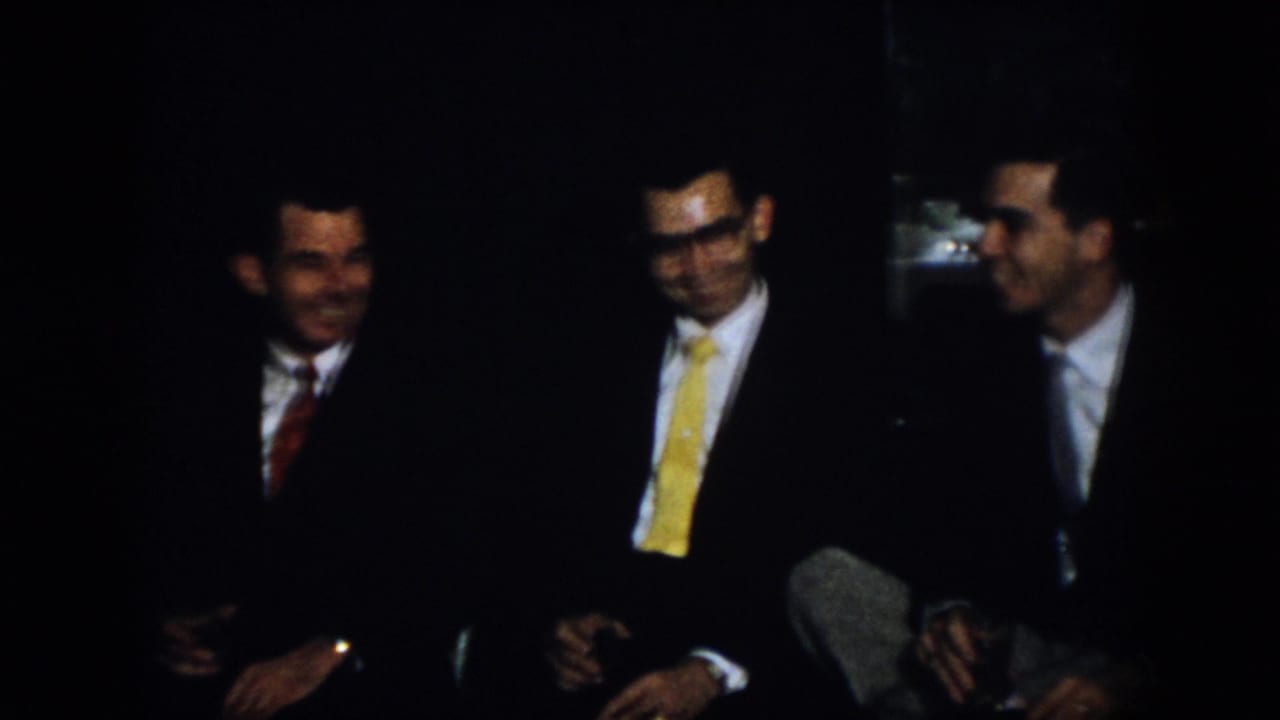 Virginia 1958 home-movie frame showing three men seated close together in a dark room, with suits, ties, smiles, and conversational body language visible.