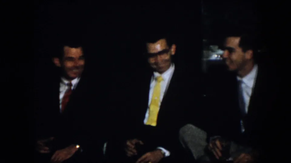 Virginia 1958 home-movie frame showing three men seated close together in a dark room, with suits, ties, smiles, and conversational body language visible.
