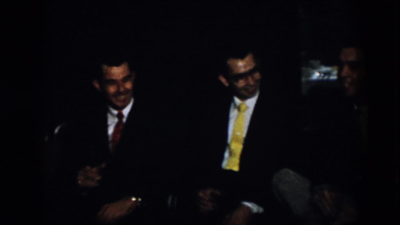 Virginia 1958 home-movie frame showing three formally dressed men seated in a shadowed interior, smiling and facing one another during conversation.