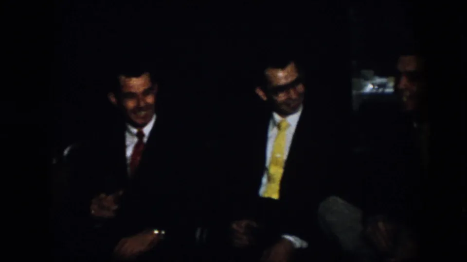 Virginia 1958 home-movie frame showing three formally dressed men seated in a shadowed interior, smiling and facing one another during conversation.
