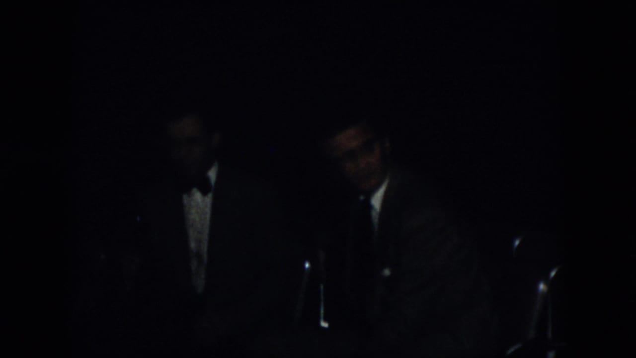 1958 Virginia 8mm frame showing men in suits near microphones under very low light at a formal indoor event.