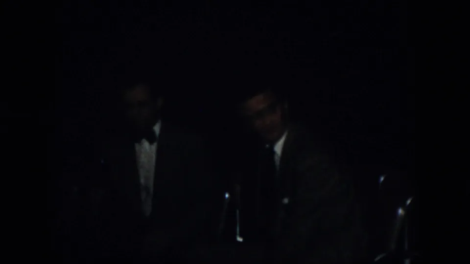 1958 Virginia 8mm frame showing men in suits near microphones under very low light at a formal indoor event.