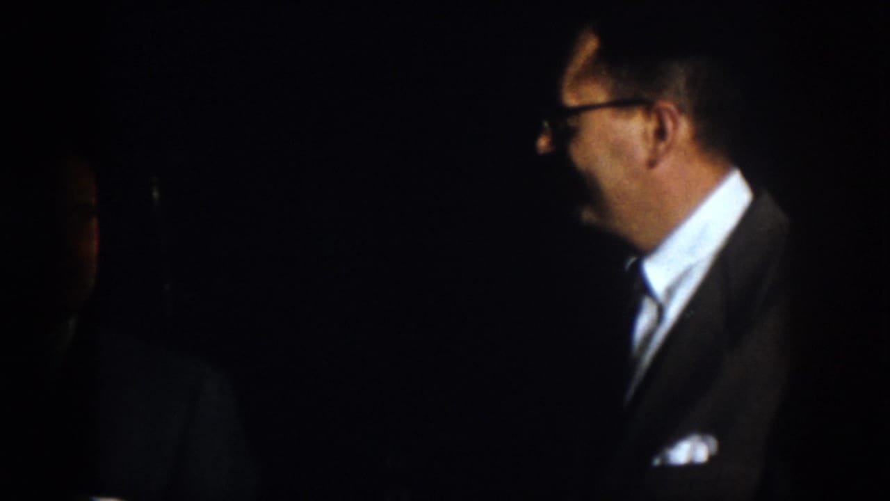 Man with glasses in a suit shown in profile against a dark background.