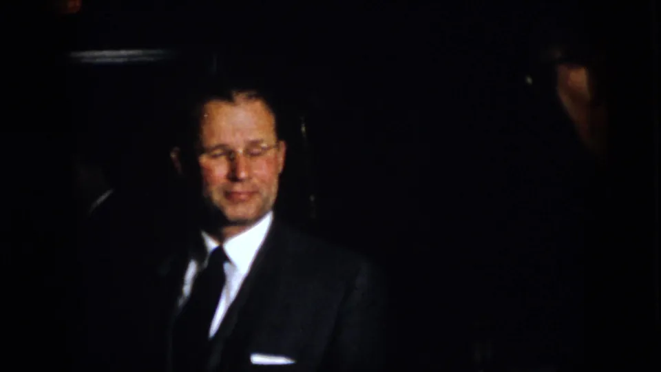 1958 Virginia 8mm frame showing a suited man smiling or reacting in a dark indoor formal setting.