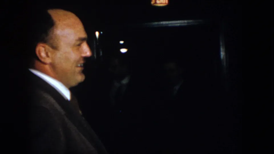 1958 Virginia frame showing a balding suited man in side profile during a low-light formal event.
