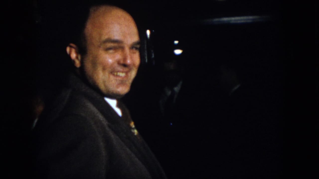 Bald man in a dark suit smiles in a dim room.