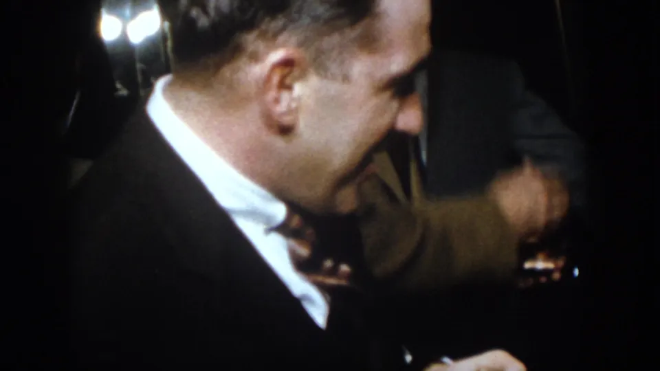 A close handheld frame shows a suited man leaning forward at the same dim 1958 Virginia social gathering, with another glass partly visible nearby.