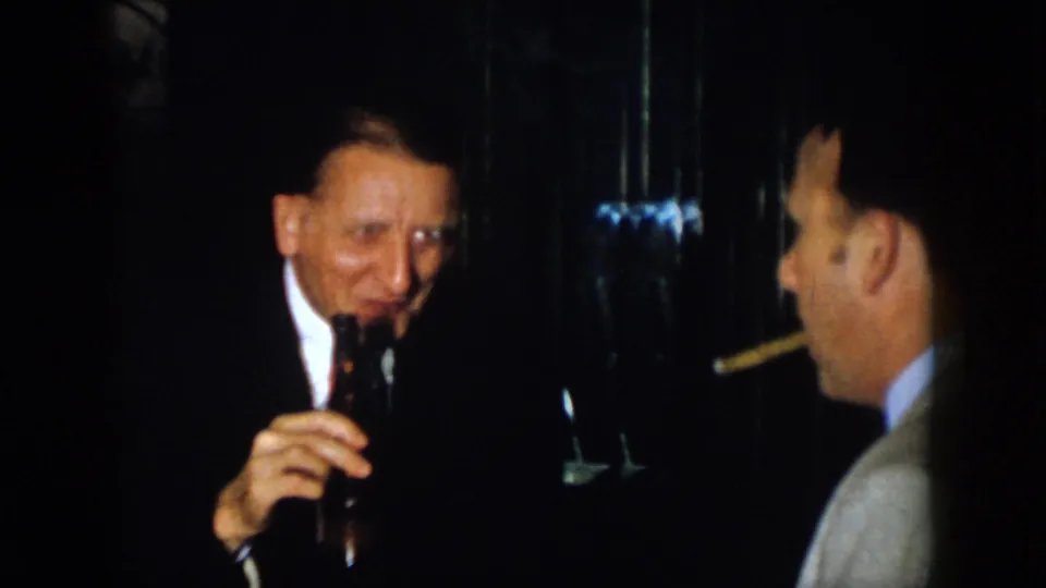 Two mature men in suits converse in a dark 1958 Virginia interior, with one holding a bottle and the other facing him with a cigar.