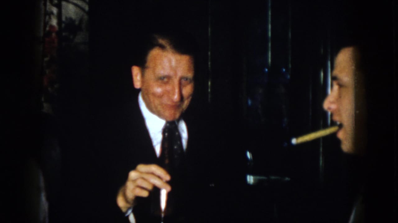 In 1958 Virginia home-movie footage, a suited man smiles toward the camera while holding a dark bottle indoors.