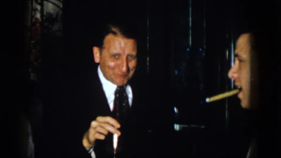 In 1958 Virginia home-movie footage, a suited man smiles toward the camera while holding a dark bottle indoors.