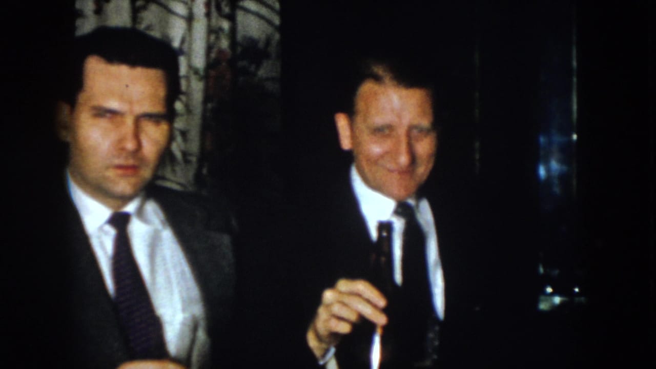 Two men in suits stand close together, with one holding a cigar.