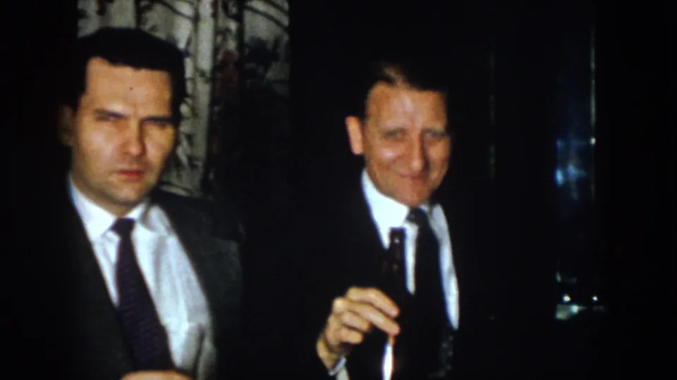 Two formally dressed men appear in a dark interior, one seen in profile with a cigar and another facing forward.