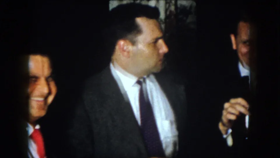 A man in a gray suit is seen in profile as other formally dressed men smile nearby in the Virginia home-movie scene.