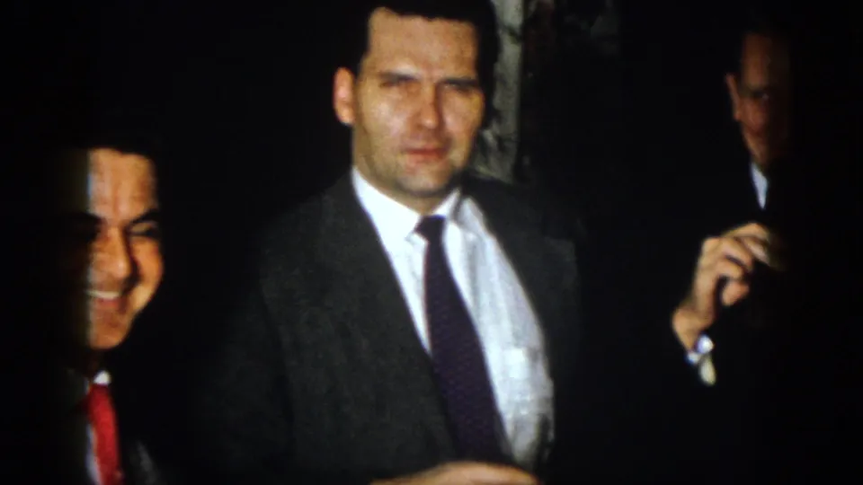A man in a gray suit faces the camera as other men smile at the edge of the dim 1958 Virginia home-movie frame.