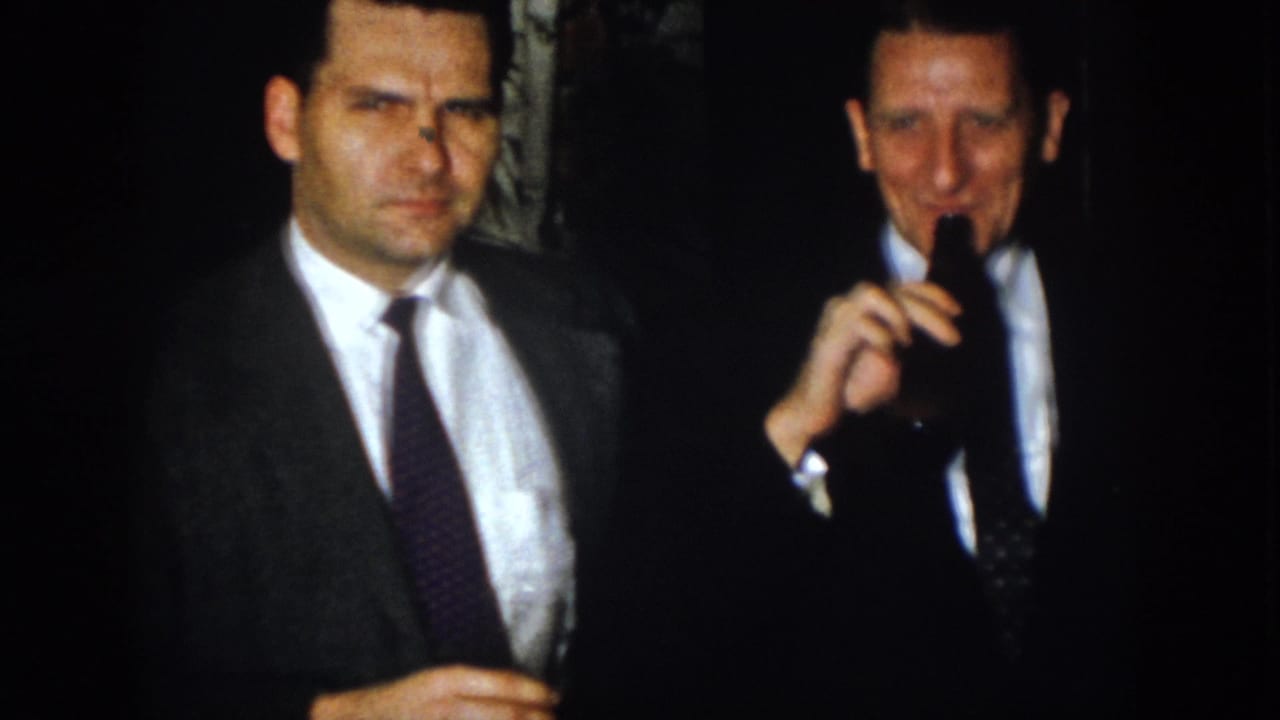 In 1958 Virginia home-movie footage, two men in suits stand at a dim nighttime gathering while one holds a dark bottle.