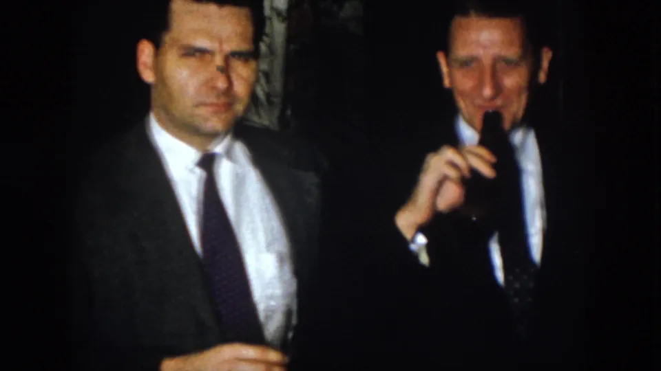 In 1958 Virginia home-movie footage, two men in suits stand at a dim nighttime gathering while one holds a dark bottle.