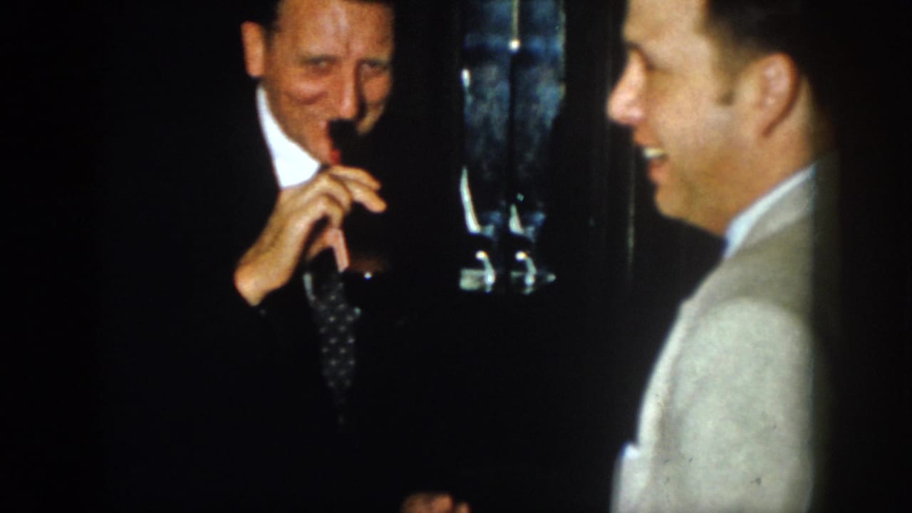 A suited man drinks from a dark bottle while another man in a light jacket smiles nearby.