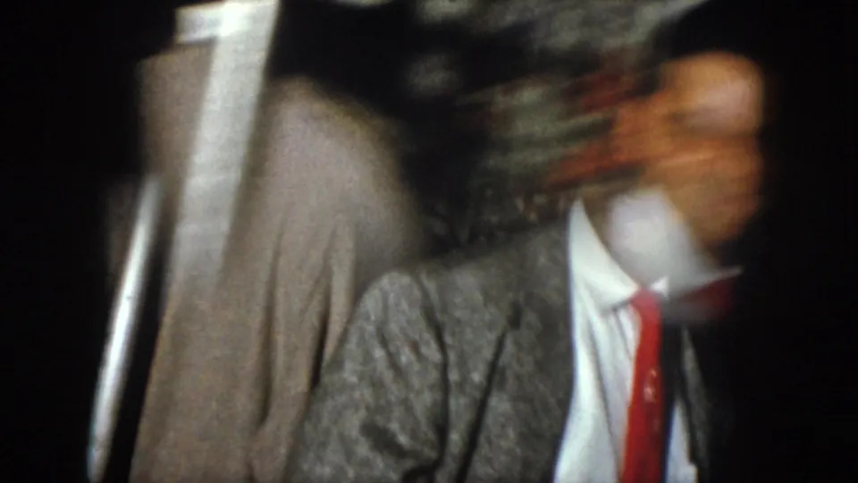 A blurred handheld frame shows suited men in motion, including a white shirt and red tie, in low light.