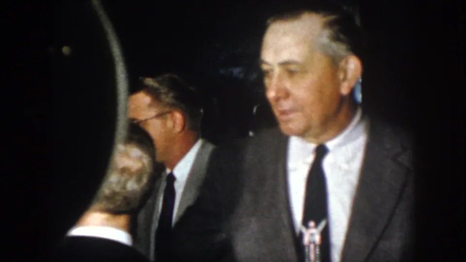 Several suited men converse in a dim social setting, with an older man in a gray suit facing toward the camera.