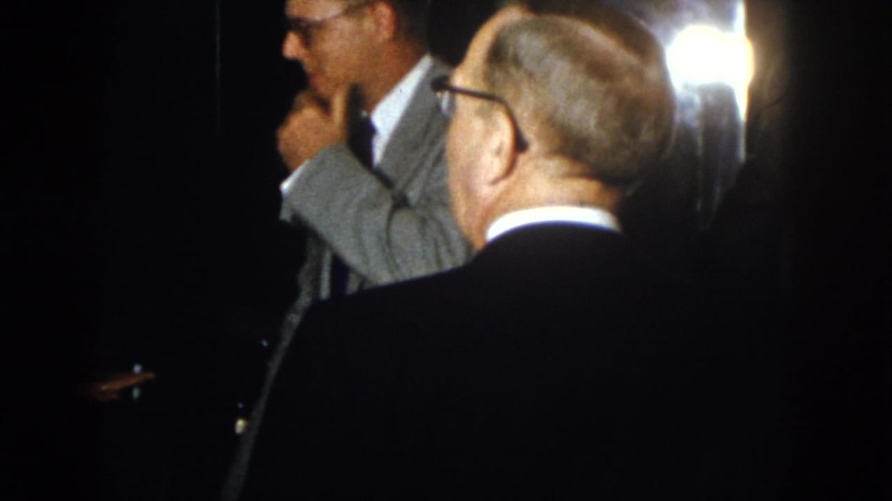 Several suited men face different directions in low light, one touching his chin.