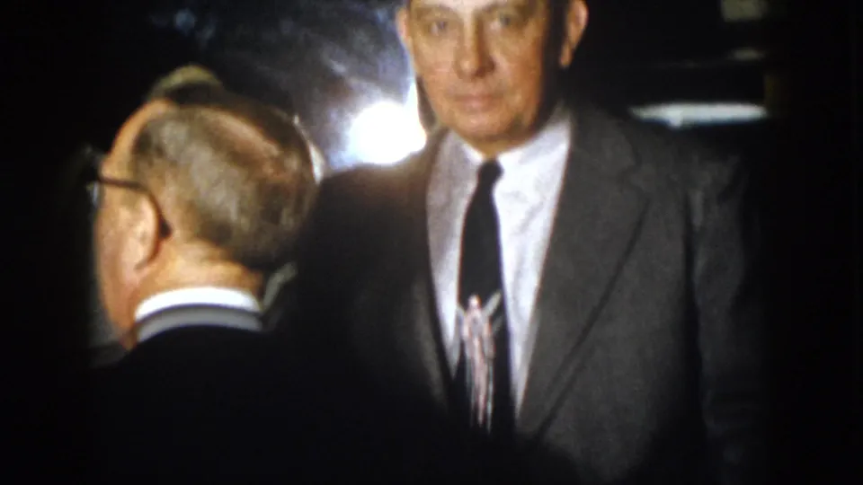 1958 Virginia home-movie still with a gray-suited man facing the camera, surrounded by other men at a night event.