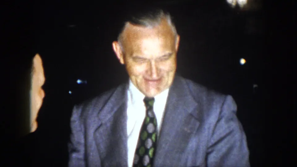 1958 Virginia frame showing an older man in a light suit and patterned tie smiling during a nighttime gathering.